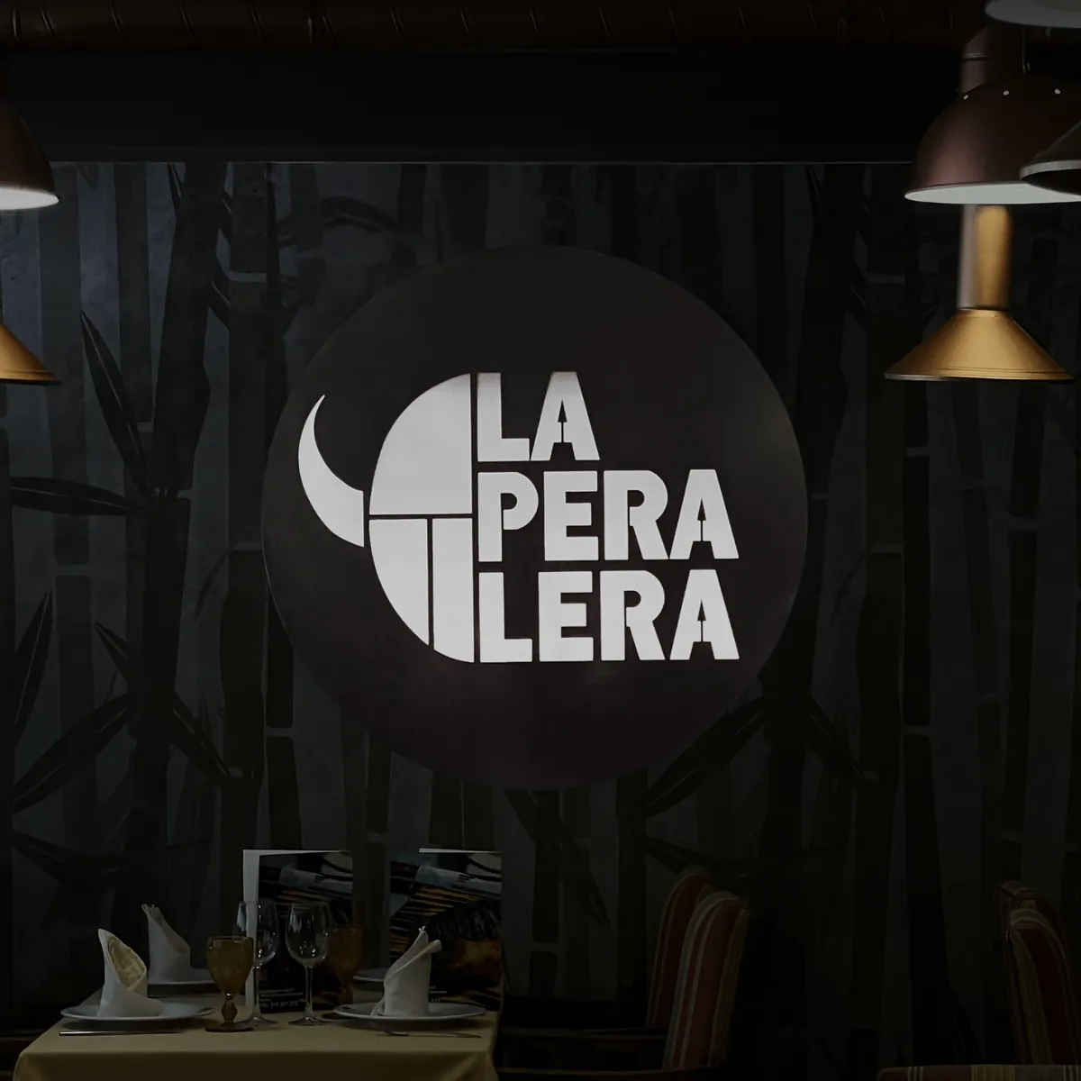 Logo de La Peralera iluminado sobre la pared de bambú del comedor